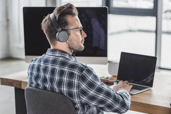 Schöner junger Programmierer mit Brille und Kopfhörer, der mit Laptop und Desktop-Computer arbeitet — Stockfoto