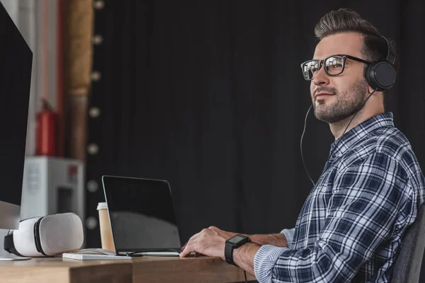 Jeune homme souriant dans les lunettes et les écouteurs de travail avec ordinateur portable — Photo de stock