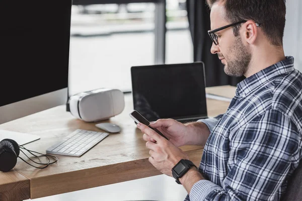 Bello giovane programmatore in occhiali utilizzando smartphone sul posto di lavoro — Foto stock