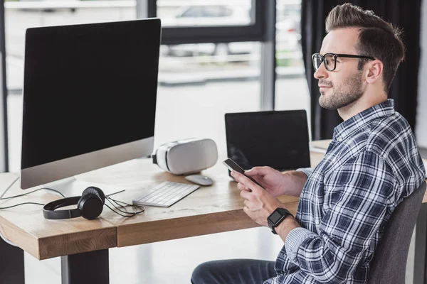 Smiling young man in eyeglasses using smartphone and looking away at workplace — Stock Photo