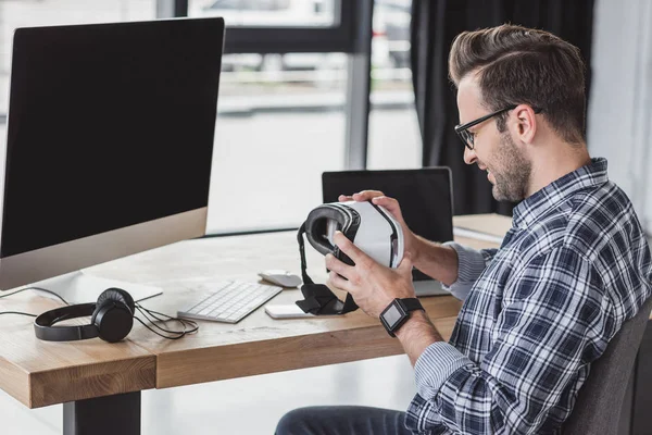 Vue latérale d'un jeune homme souriant portant des lunettes tenant un casque de réalité virtuelle sur le lieu de travail — Photo de stock