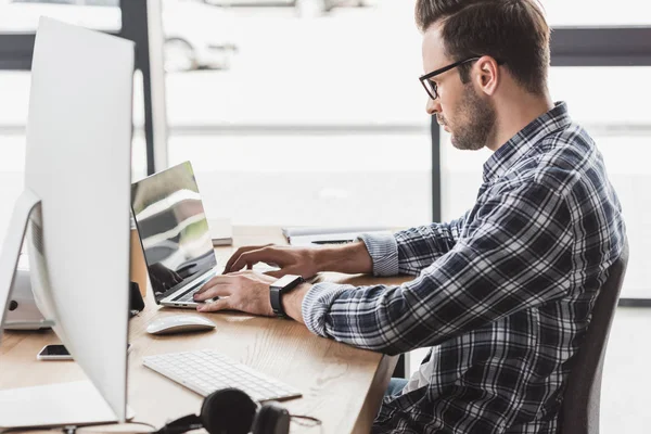 Visão lateral de bonito jovem programador em óculos trabalhando com laptop e computador desktop — Fotografia de Stock