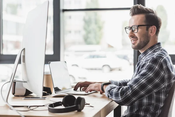 Joven sonriente en gafas con computadora portátil y computadora de escritorio en el lugar de trabajo - foto de stock