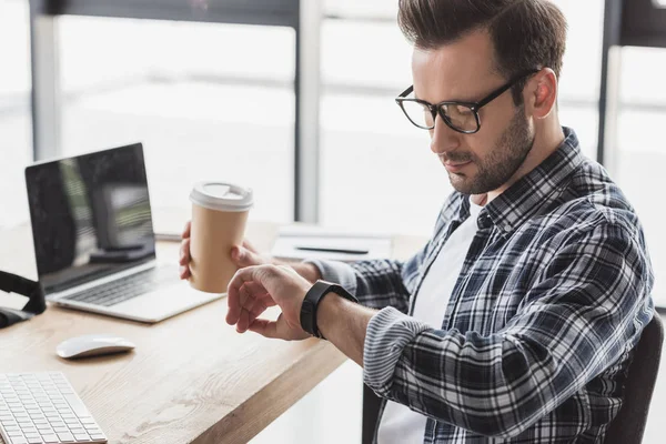 Junger Mann mit Brille hält Coffee to go und checkt Smartwatch am Arbeitsplatz — Stockfoto
