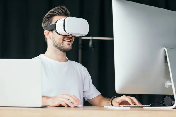 Bonito sorrindo jovem no fone de ouvido realidade virtual usando computador desktop e laptop — Fotografia de Stock