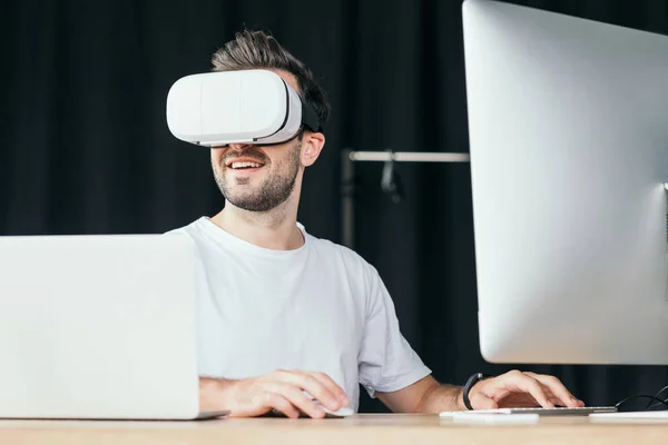 Smiling young man in virtual reality headset using desktop computer and laptop — Stock Photo