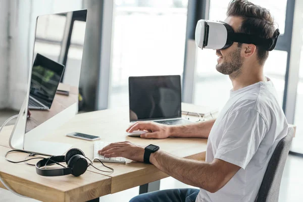 Jovem sorridente em fone de ouvido de realidade virtual usando computador desktop no local de trabalho — Fotografia de Stock