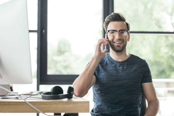 Joven guapo en anteojos hablando por teléfono inteligente y sonriendo a la cámara mientras está sentado en el lugar de trabajo - foto de stock