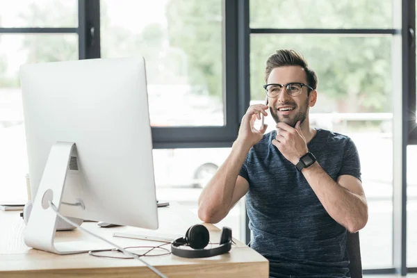 Guapo joven sonriente en gafas que habla por teléfono inteligente mientras trabaja con la computadora de escritorio - foto de stock