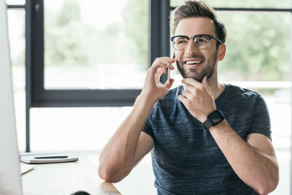 Bonito sorrindo jovem em óculos falando por smartphone no local de trabalho — Fotografia de Stock