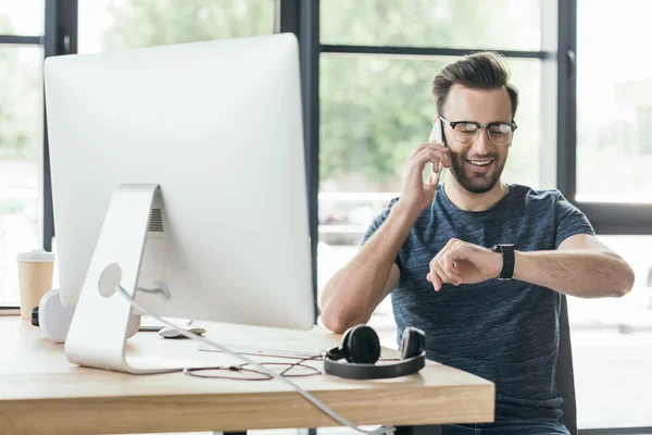 Jovem sorridente em óculos verificando smartwatch e falando por smartphone no local de trabalho — Fotografia de Stock