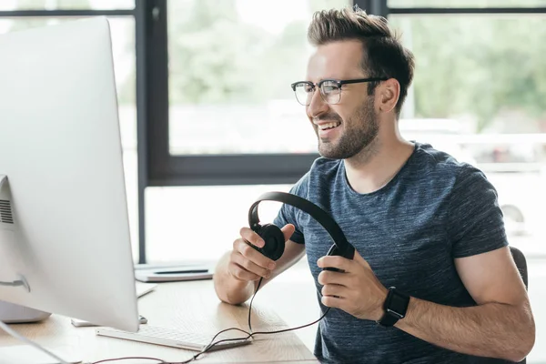 Bonito sorrindo homem em óculos segurando fones de ouvido e usando computador desktop — Fotografia de Stock