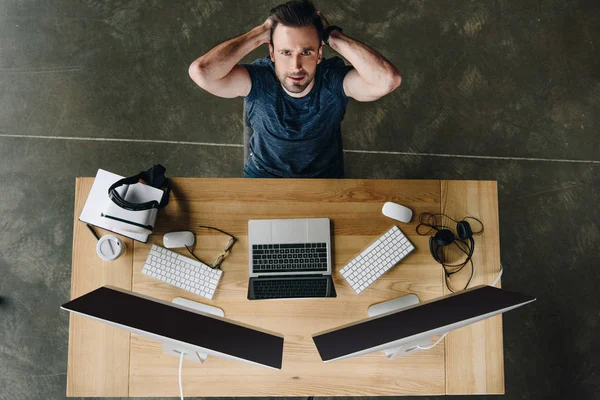 Красивый молодой человек с руками за головой смотрит в камеру, сидя на рабочем месте с компьютерами — стоковое фото