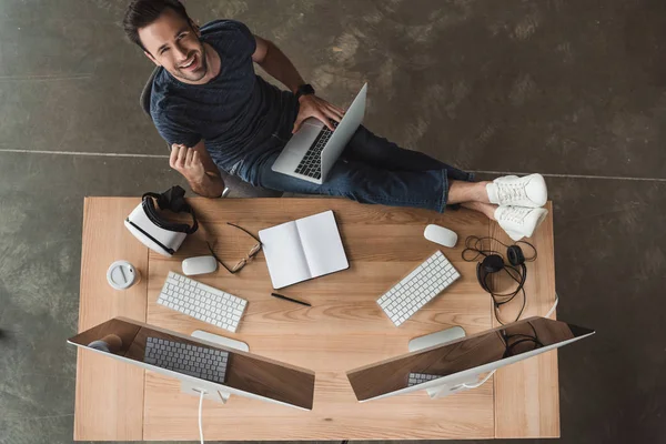 Overhead view of handsome young programmer using laptop and smiling at camera — Stock Photo