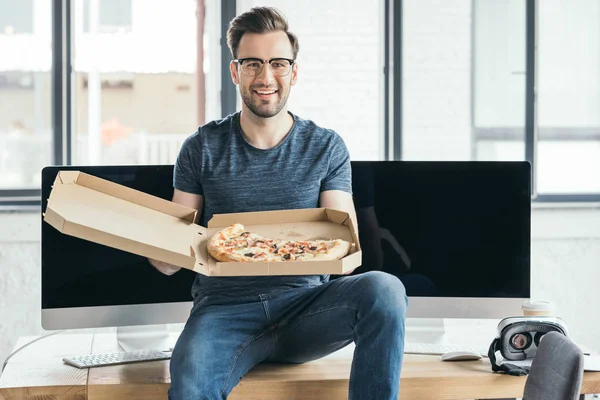 Bonito jovem programador no óculos segurando pizza e sorrindo para a câmera — Fotografia de Stock