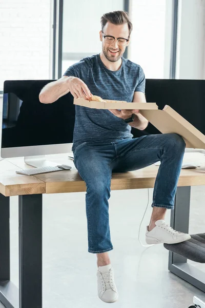 Beau jeune programmeur souriant dans des lunettes manger de la pizza sur le lieu de travail — Photo de stock