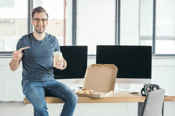 Sonriente joven programador en anteojos sosteniendo taza de papel y comiendo pizza en el lugar de trabajo - foto de stock