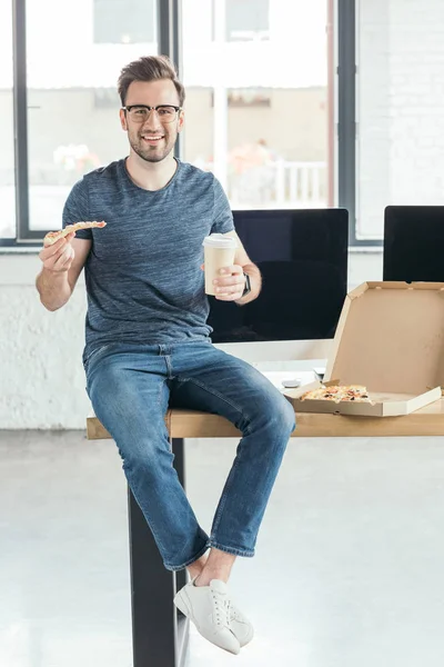 Schöner lächelnder junger Programmierer mit Brille, Pappbecher in der Hand und Pizza essen am Arbeitsplatz — Stockfoto