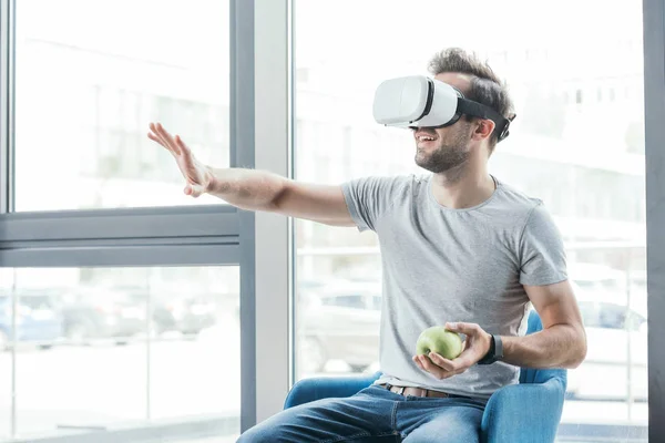 Feliz joven en realidad virtual auricular sosteniendo manzana y sentado en la silla - foto de stock