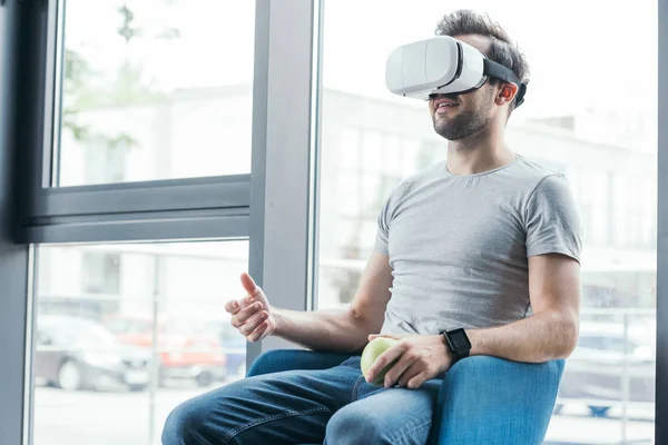 Улыбающийся молодой человек в наушниках виртуальной реальности держит яблоко и сидит на стуле — стоковое фото