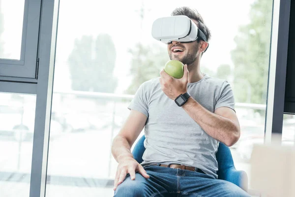 Sorridente jovem segurando maçã verde e usando fone de ouvido realidade virtual — Fotografia de Stock