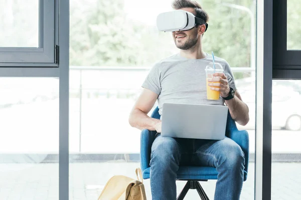 Sorrindo jovem segurando copo de plástico com suco fresco e usando fone de ouvido realidade virtual com laptop — Fotografia de Stock