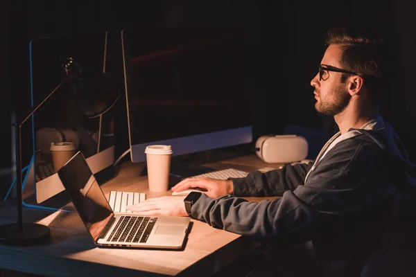 Seitenansicht eines jungen Programmierers in Brille, der nachts mit Laptop und Desktop-Computer arbeitet — Stockfoto