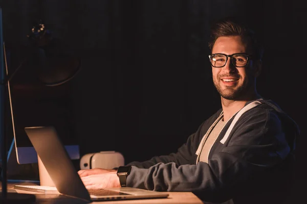 Schöner junger Programmierer mit Brille lächelt in die Kamera, während er nachts mit Laptop und Desktop-Computer arbeitet — Stockfoto