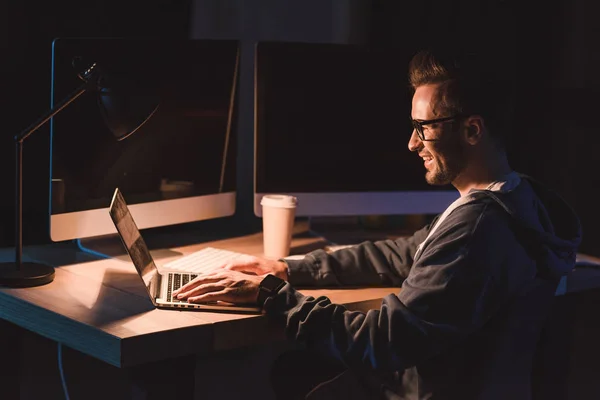 Sonriente joven programador en gafas usando portátil mientras trabaja por la noche - foto de stock