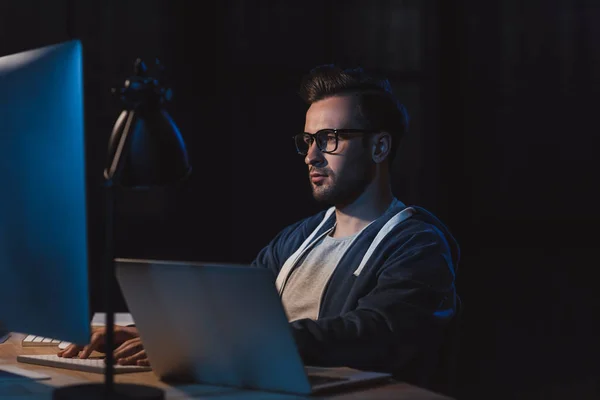 Молодий програміст в окулярах працює з настільним комп'ютером і ноутбуком вночі — стокове фото