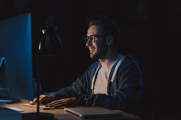 Sonriente joven programador en anteojos trabajando con ordenador de sobremesa en la noche - foto de stock