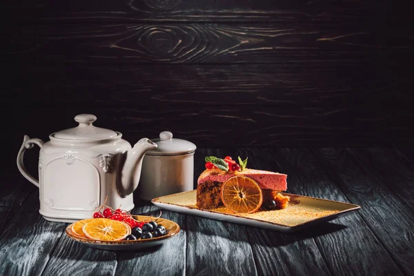 Carrot cake with berry filling on plate, orange slices, blueberries and cranberries on saucer near teapot on wooden table — Stock Photo