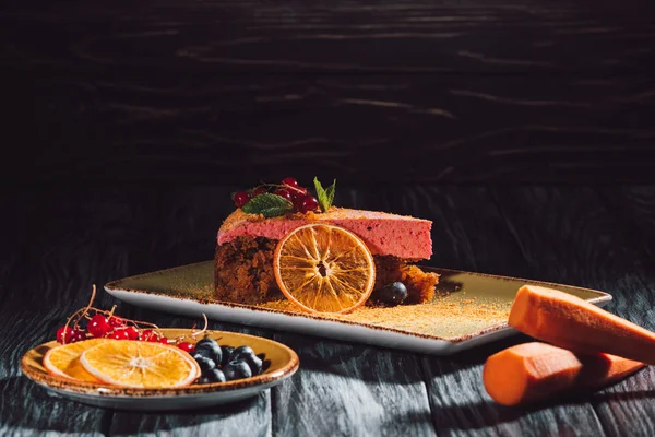 Close up view of carrot cake with mint and berry filling, carrots, orange slices and berries on saucer on wooden table — Stock Photo