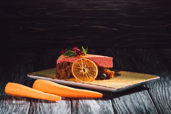 Vista da vicino della torta di carote con ripieno di bacche, menta, fetta d'arancia sul piatto vicino alle carote sul tavolo di legno — Foto stock