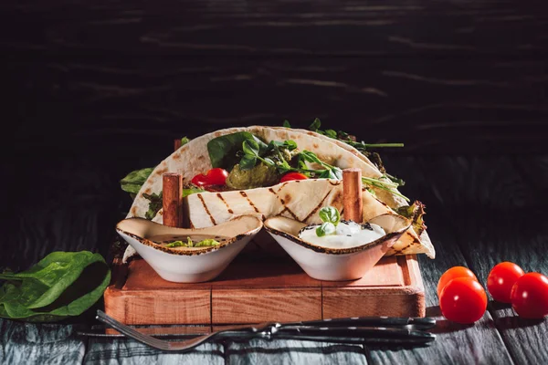 Close up view of tortillas with falafel, cherry tomatoes and germinated seeds of sunflower on wooden tray with sauces near fork and knife on wooden table — Stock Photo