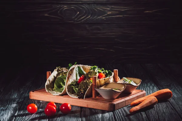 Selective focus of tortillas with falafel, cherry tomatoes and germinated seeds of sunflower on wooden tray with sauces near carrots — Stock Photo