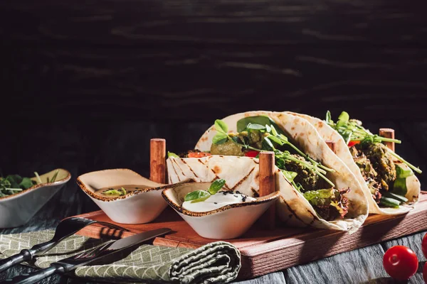 Composizione alimentare di tortillas con falafel, pomodorini e semi germinati di girasole su vassoio di legno con salse vicino a forchetta e coltello sul tavolo — Foto stock