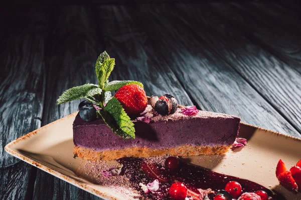 Vista de cerca de pastel de arándanos con fresas, menta y pétalos de viola en el plato en la mesa de madera - foto de stock