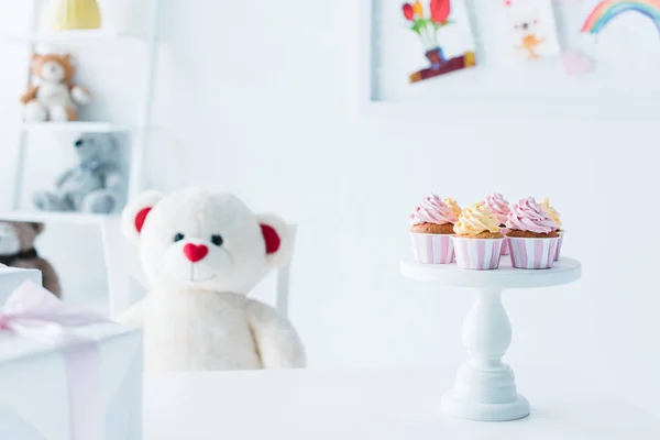 Ursinho de pelúcia sentado à mesa com cupcakes em stand — Fotografia de Stock