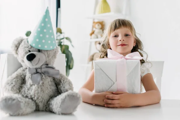 Happy birthday kid sitting with present at table with teddy bear in cone — Stock Photo