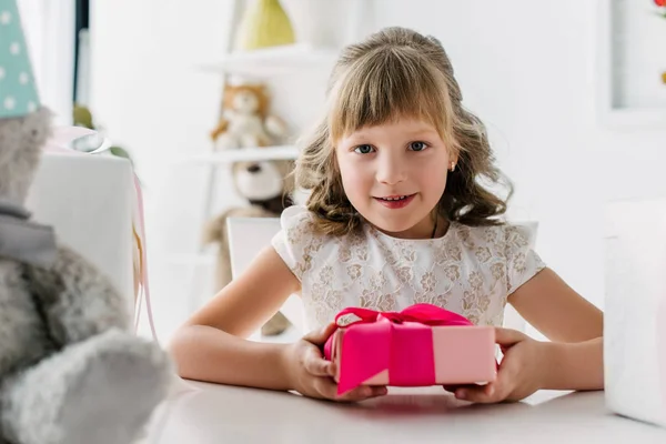 Niño de cumpleaños sonriente sosteniendo caja de regalo y mirando a la cámara en la mesa - foto de stock