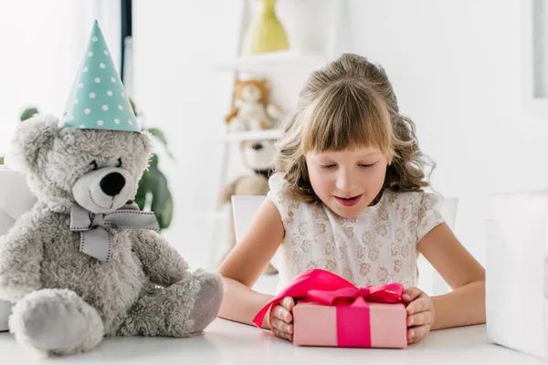 Bambino di compleanno sorridente guardando scatola regalo mentre seduto a tavola con orsacchiotto in cono — Foto stock