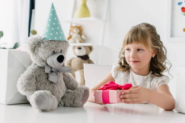 Cute little kid giving gift box to teddy bear in cone at table — Stock Photo