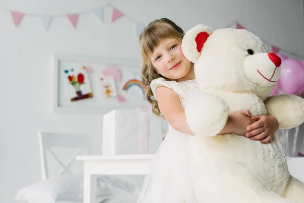 Happy birthday kid in white dress embracing teddy bear — Stock Photo