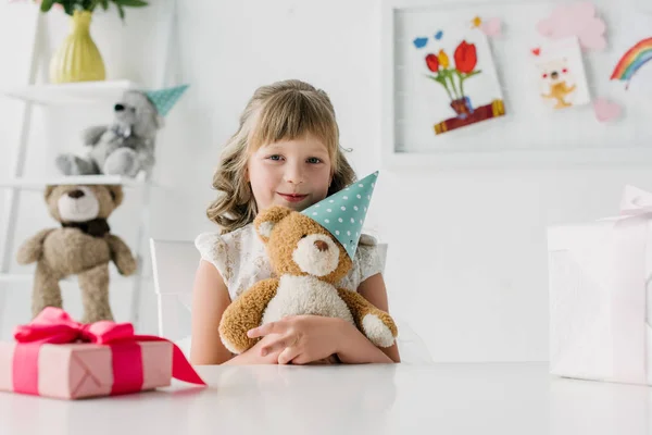 Niedliches Geburtstagskind hält Teddybär in Kegel am Tisch mit Geschenkbox — Stockfoto