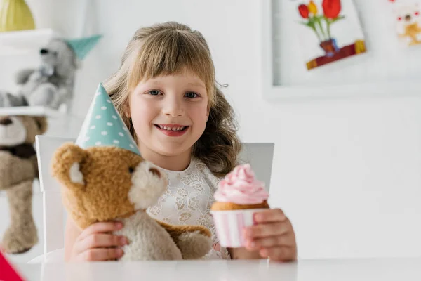 Smiling birthday kid showing cupcake and holding teddy bear in cone at table — Stock Photo