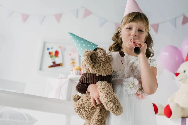 Happy birthday child in cone blowing in party horn and holding teddy bear — Stock Photo