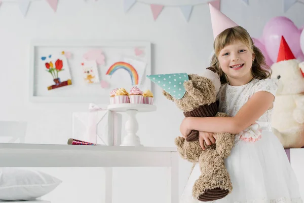 Niño de cumpleaños sonriente sosteniendo oso de peluche en cono - foto de stock