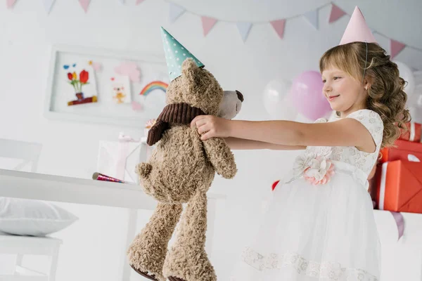 Vista lateral del niño de cumpleaños sonriente sosteniendo oso de peluche en cono - foto de stock