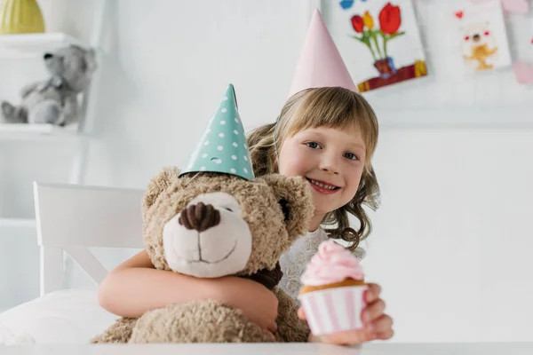 Sourire enfant anniversaire tenant ours en peluche et donner cupcake — Photo de stock